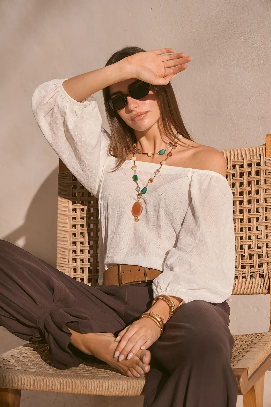 Modelo con blusa blanca de hombros descubiertos y pantalón chocolate, sentada al sol